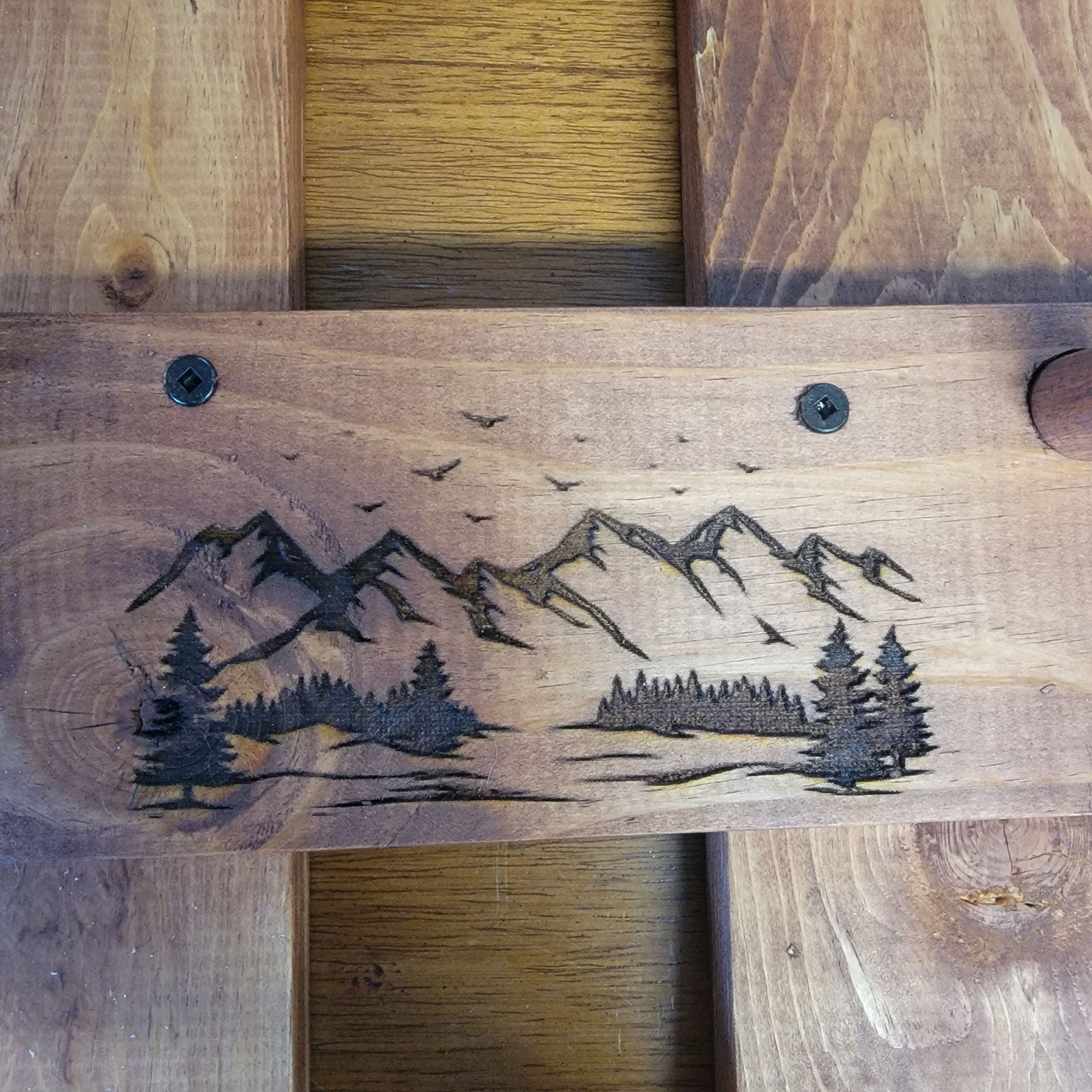 Wood plank with a mountain and tree landscape design, birds in the sky, and two screws.