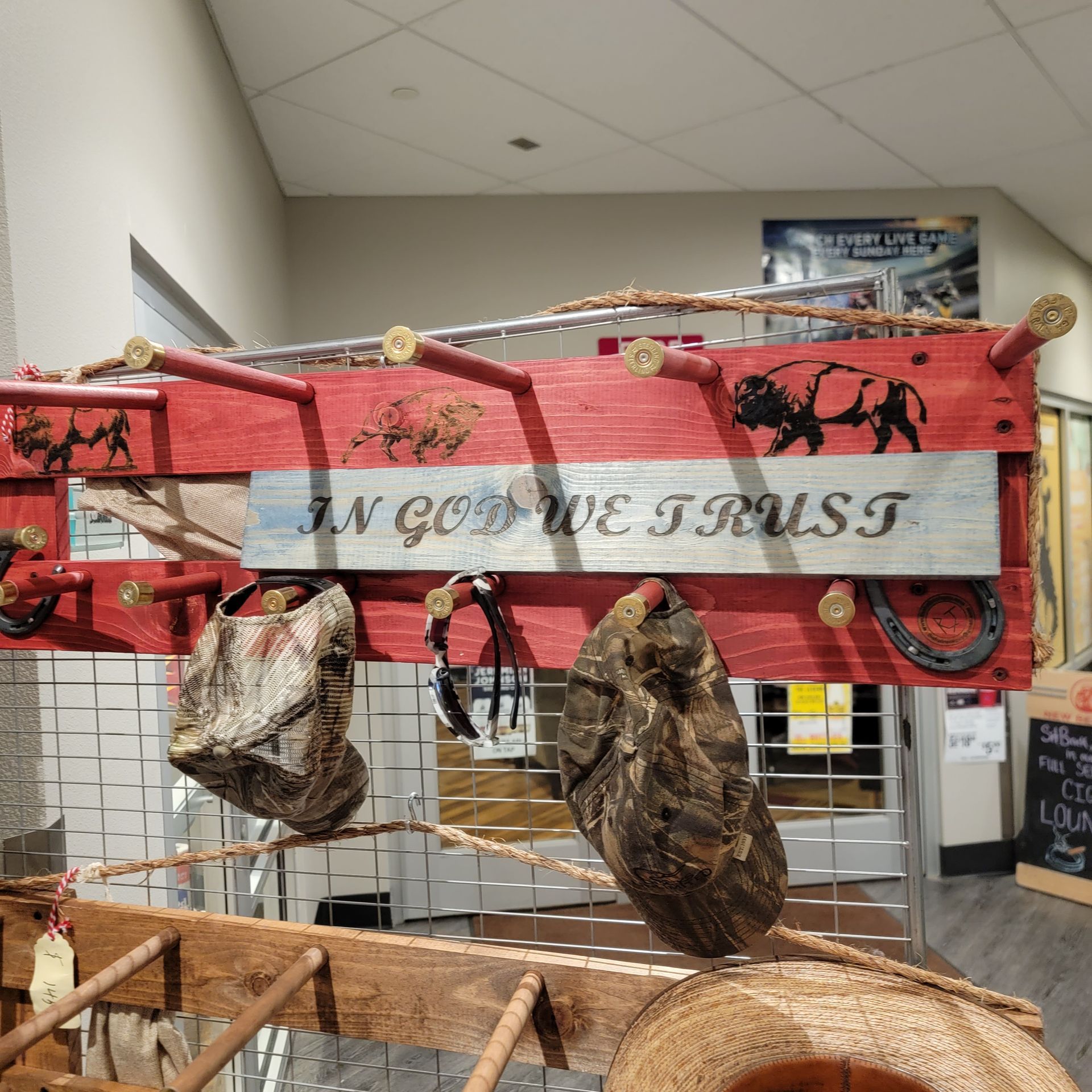 Rustic red wooden wall rack with “IN GOD WE TRUST” text and hanging items like bags, hats, and a decorative rock.