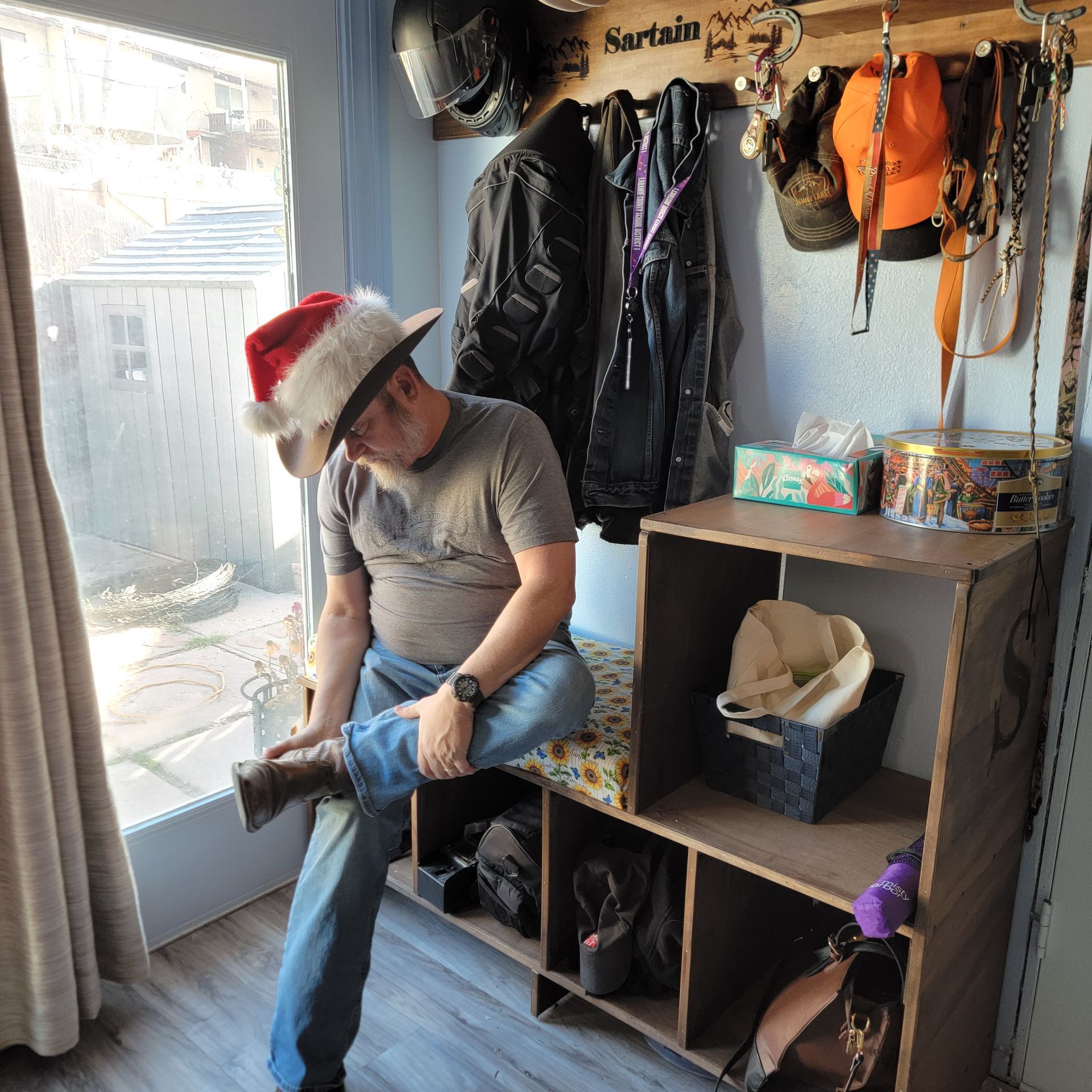 Man in Santa hat putting on a boot, sitting on a wooden bench with storage compartments, coats, and hats.