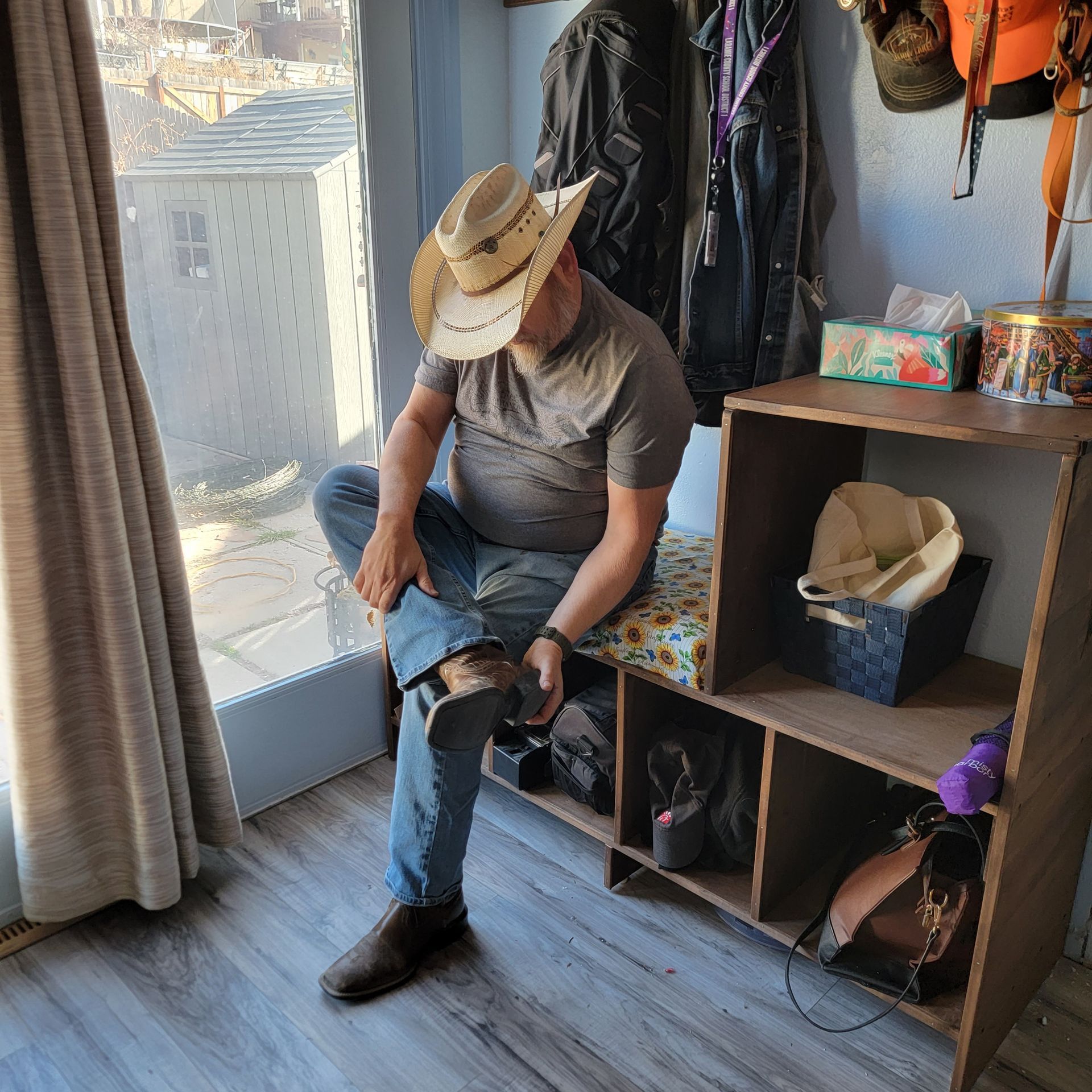 Man in a cowboy hat and jeans sits on a wooden bench, putting on a boot. Inside a house.