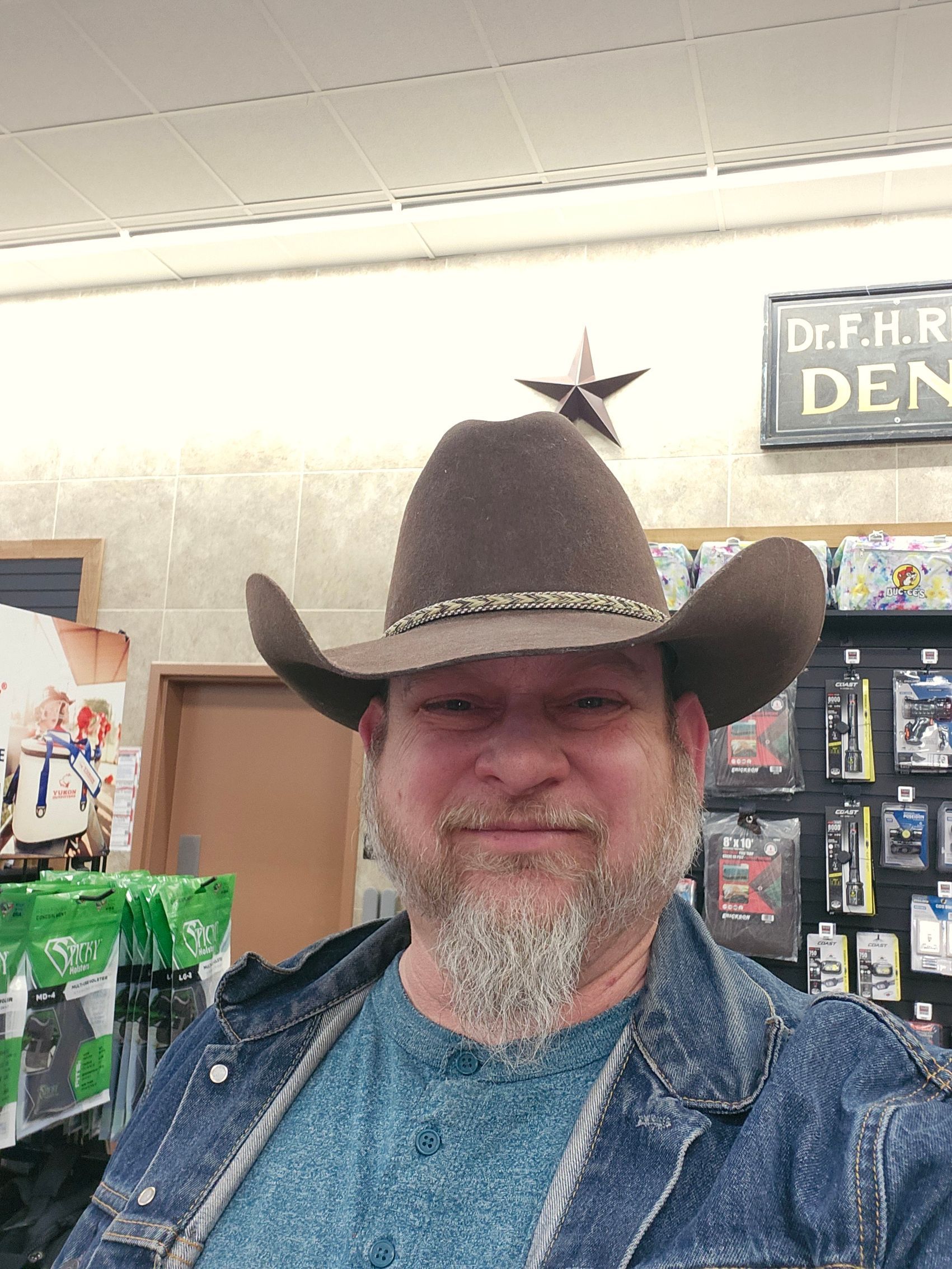 Man wearing a cowboy hat and denim jacket, smiling indoors near store shelves.
