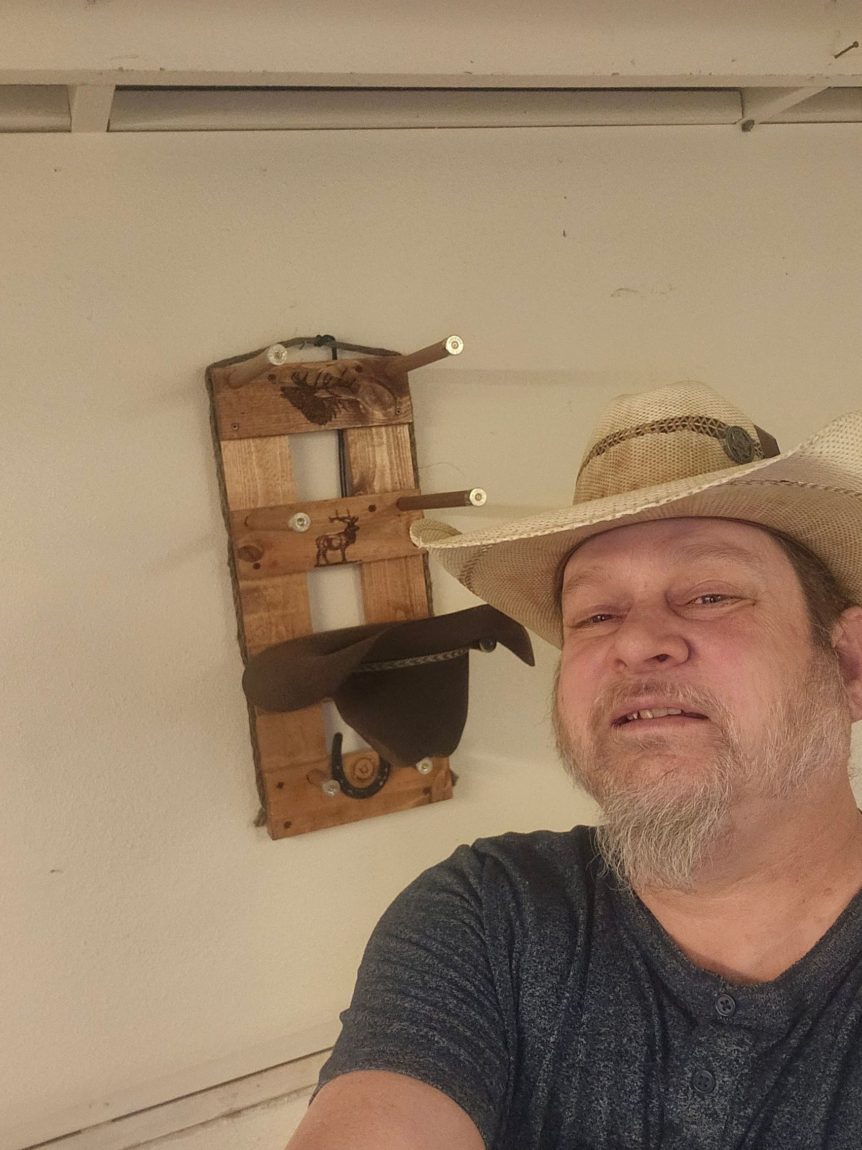 Man wearing straw hat, smiling, with hat rack on wall behind him.