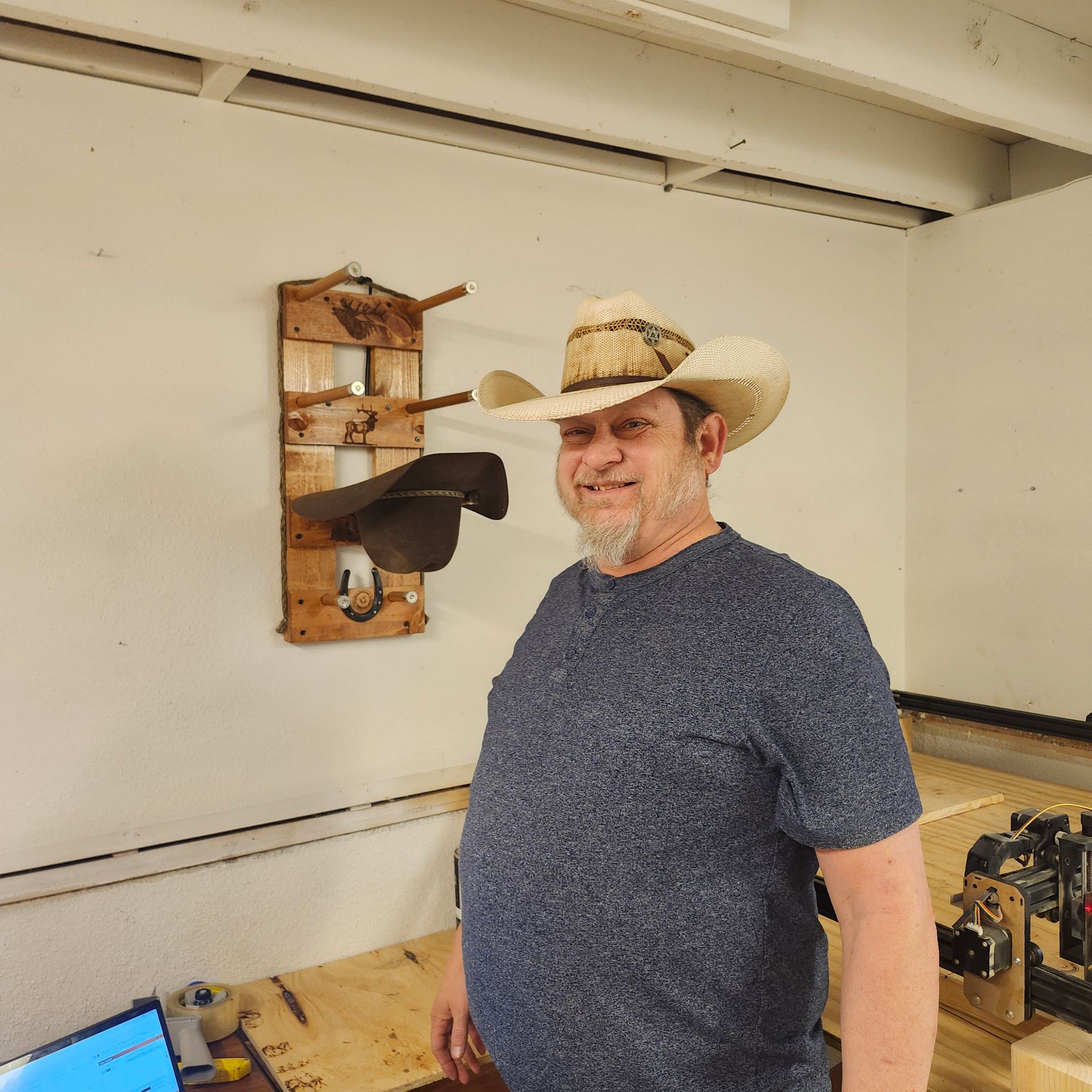 Man in cowboy hat smiles, standing by wooden saddle rack and table in a workshop.