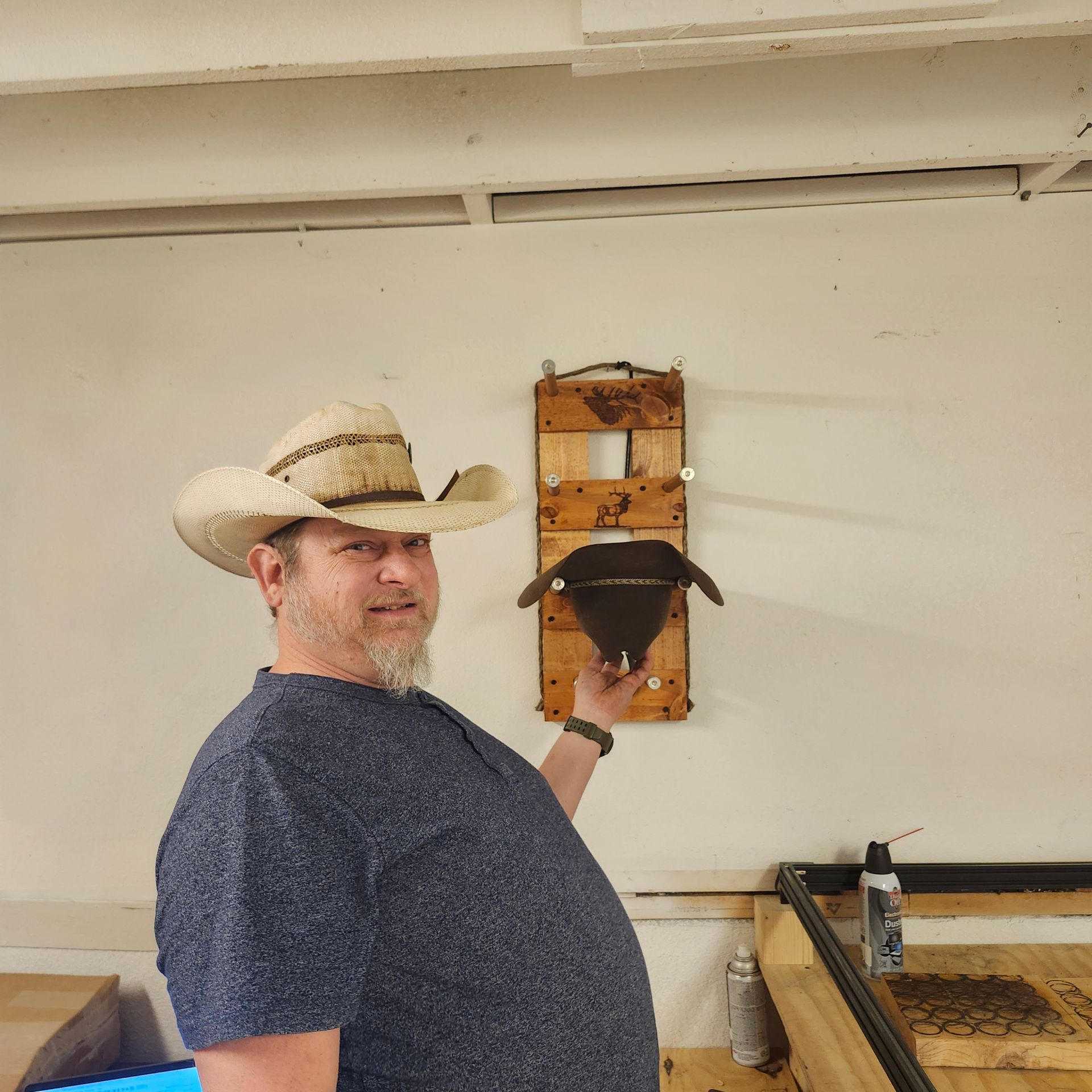 Man in cowboy hat holding up a leather mask, standing in front of wooden wall mount.