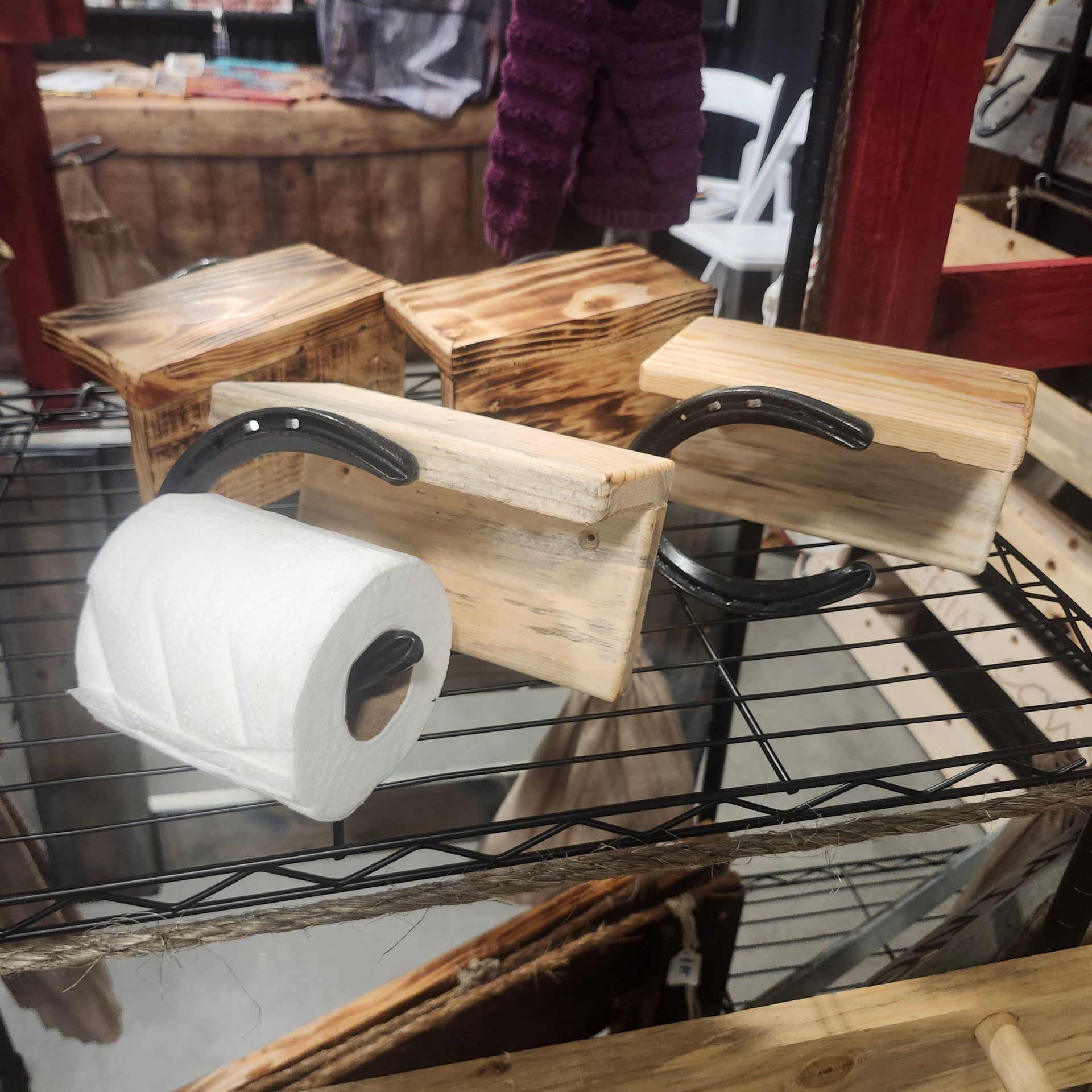 Wooden toilet paper holders with horseshoe accents displayed on a metal shelf. A roll of toilet paper rests in one.