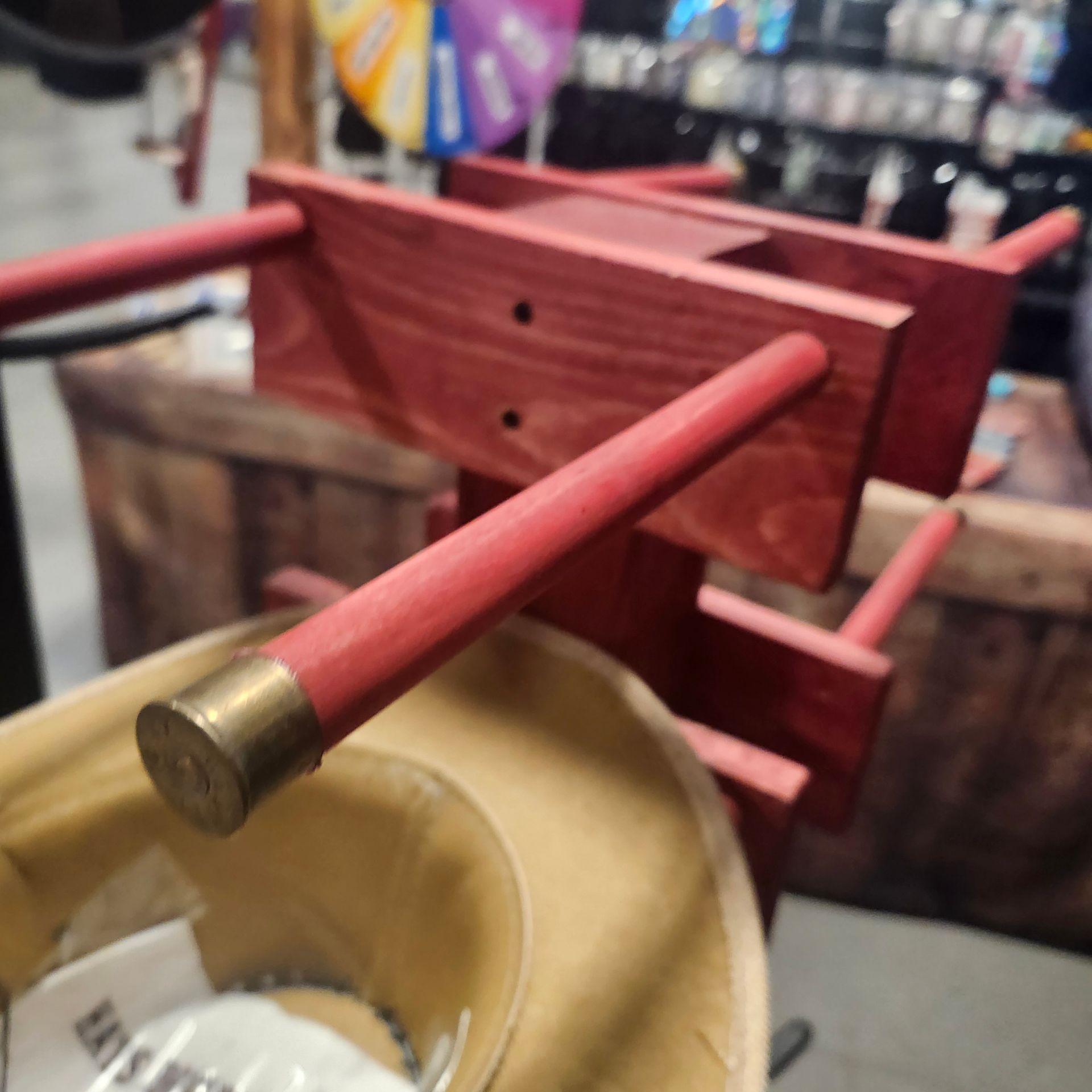 Red wooden wine rack with brass-capped pegs, resting on a hat. Retail setting.