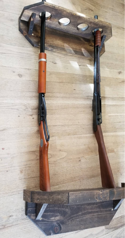 Two rifles in a wooden wall-mounted rack. Dark wood rack, brown and black rifles. Light wood wall background.