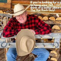Man in plaid shirt and cowboy hat, holding a tan hat, near a wooden hat rack.