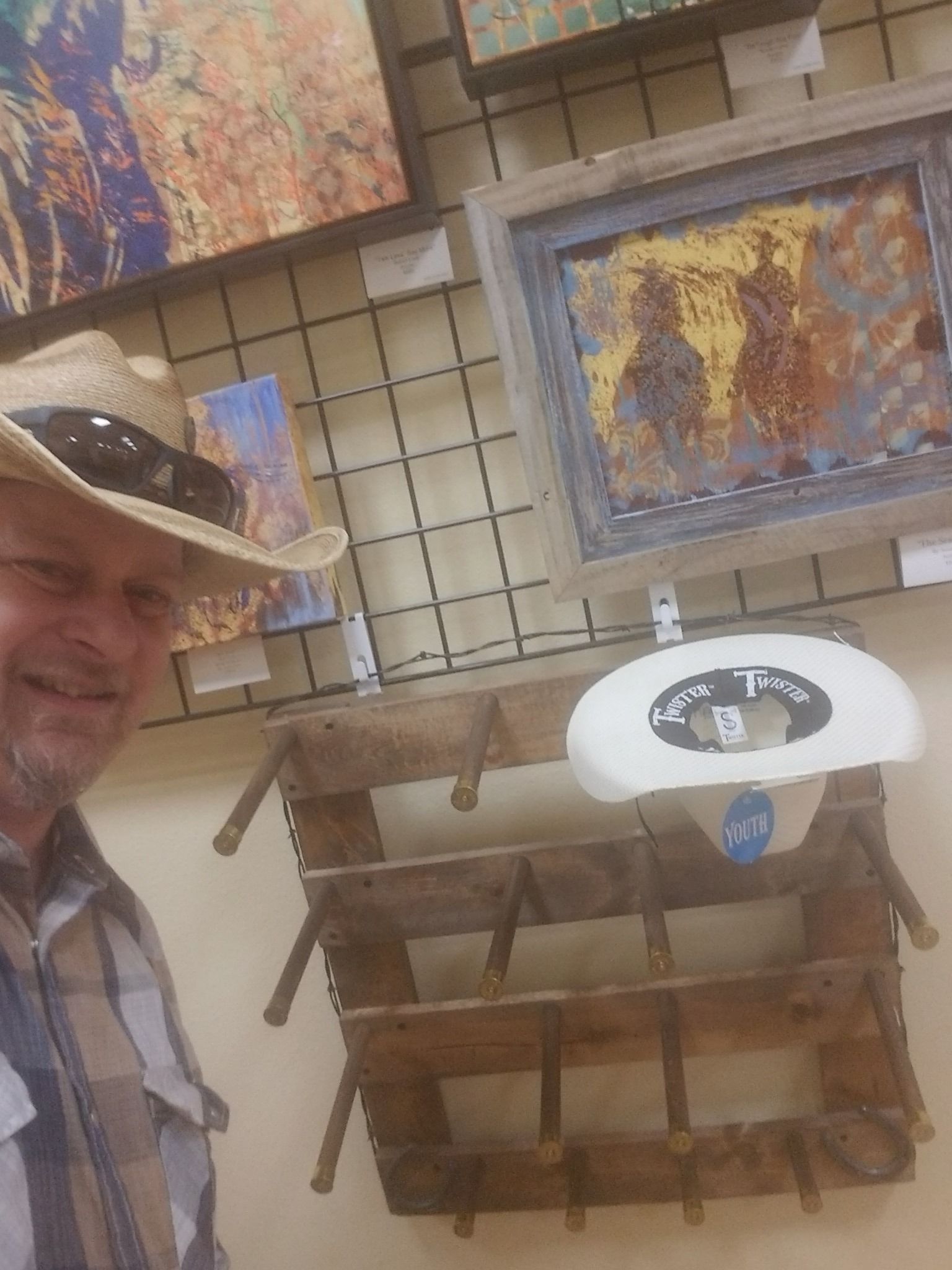 Man in hat smiles near art display; paintings on wall. Rustic shelf, white light.