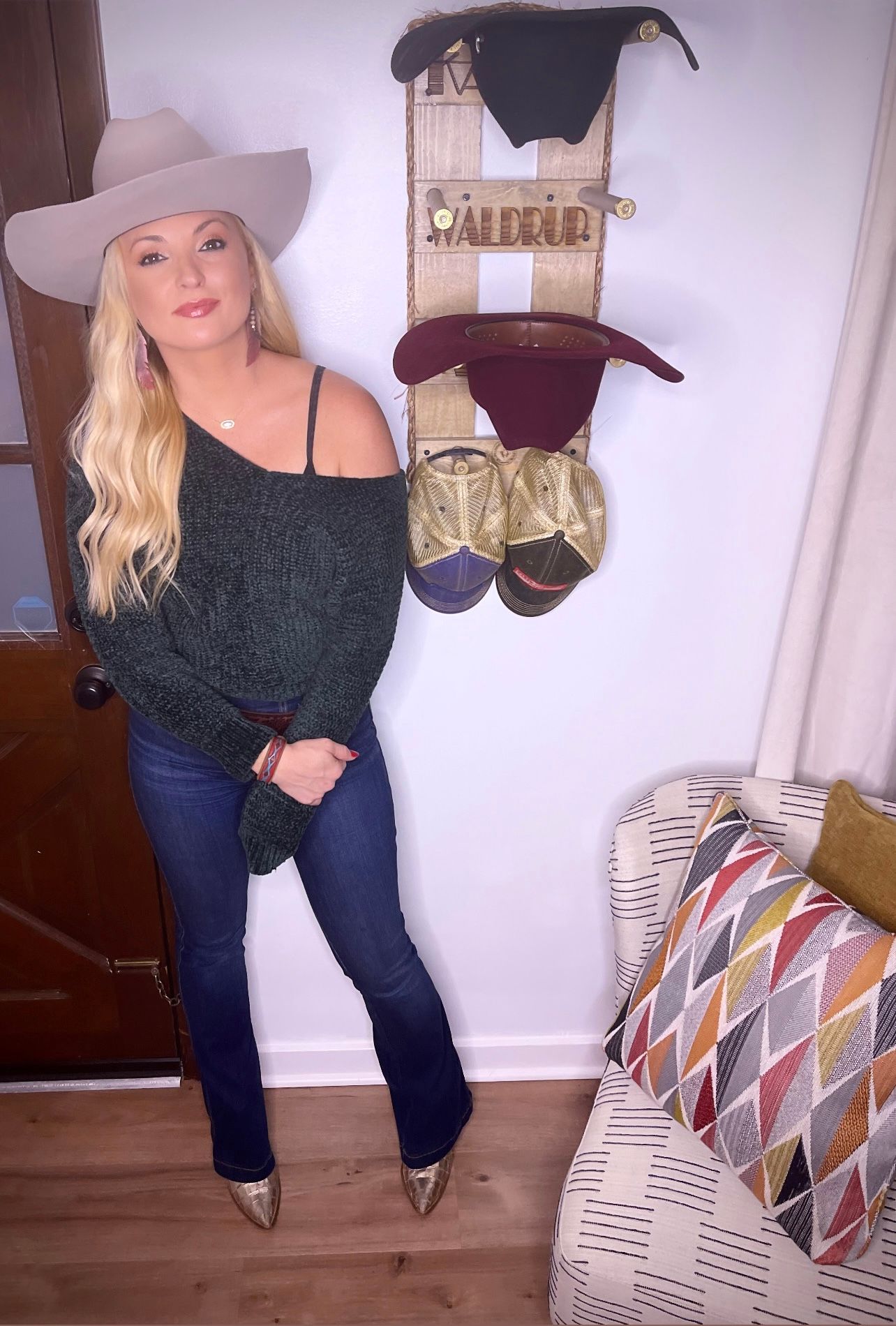 Woman in cowboy hat and off-shoulder sweater, jeans, and heels, standing near a wall-mounted hat rack, next to a chair with pillows.