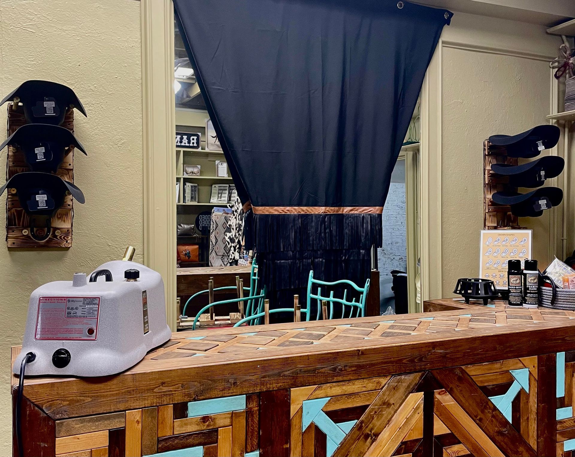 Shop interior: Wooden counter with a machine. Hats mounted on walls. Black curtain.