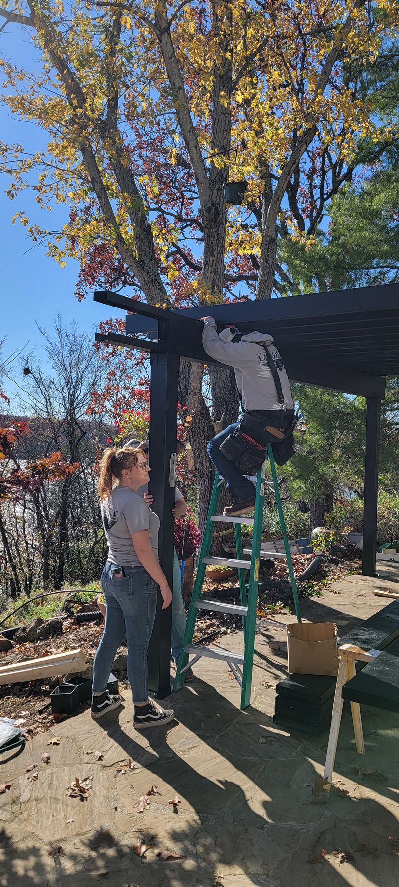 Two people beside a green ladder under a wooden pergola, one standing on top among autumn trees.