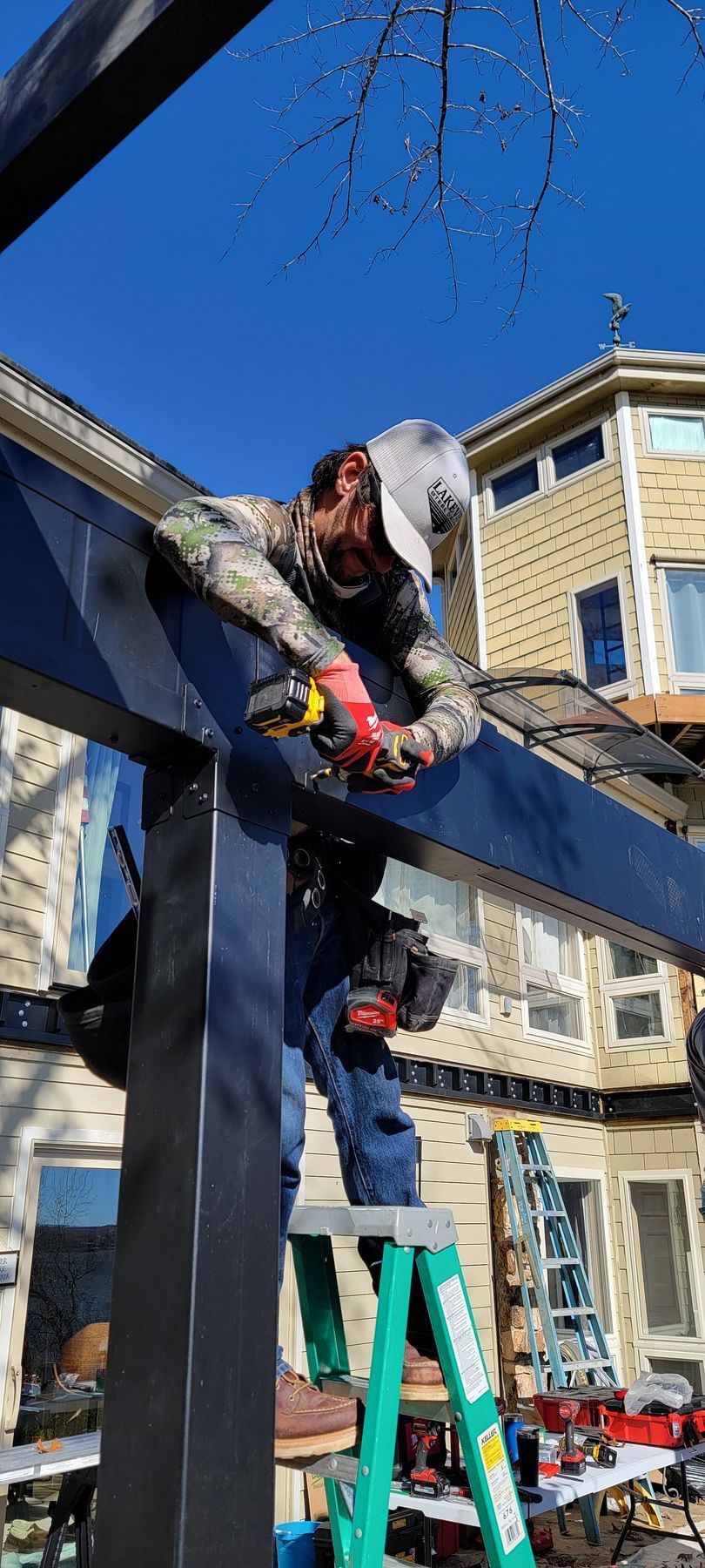 Worker on a ladder installing a beam on a blue wooden structure in front of a house.