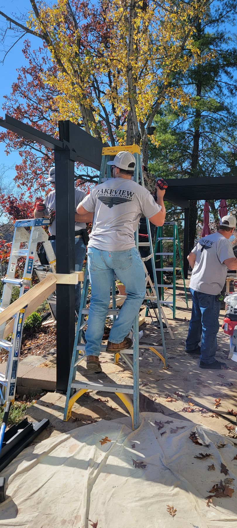 Person on a ladder working on a black post outdoors in a sunlit yard with others nearby