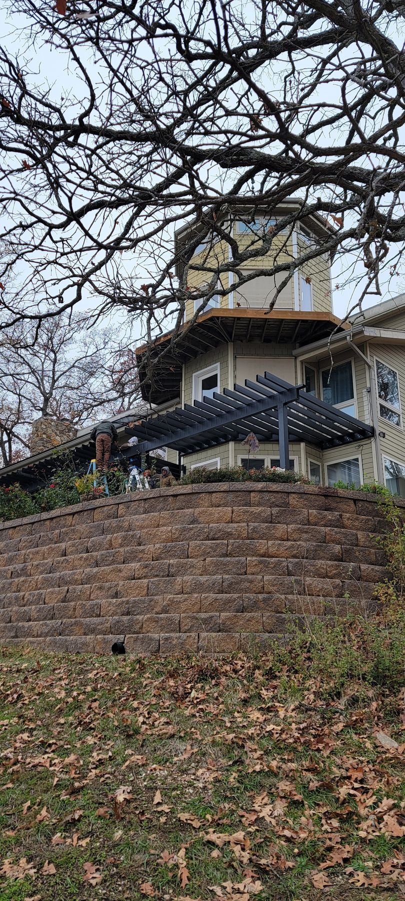 Brick retaining wall with leaf-covered ground and a house with porch and tower behind bare tree branches