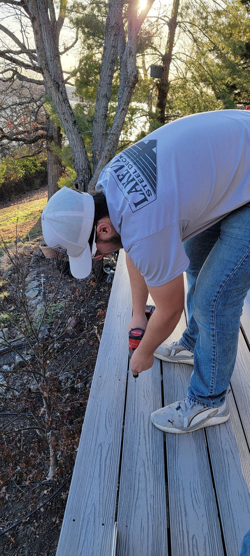 Person in gray shirt and cap bending over a wooden deck rail outdoors among trees.