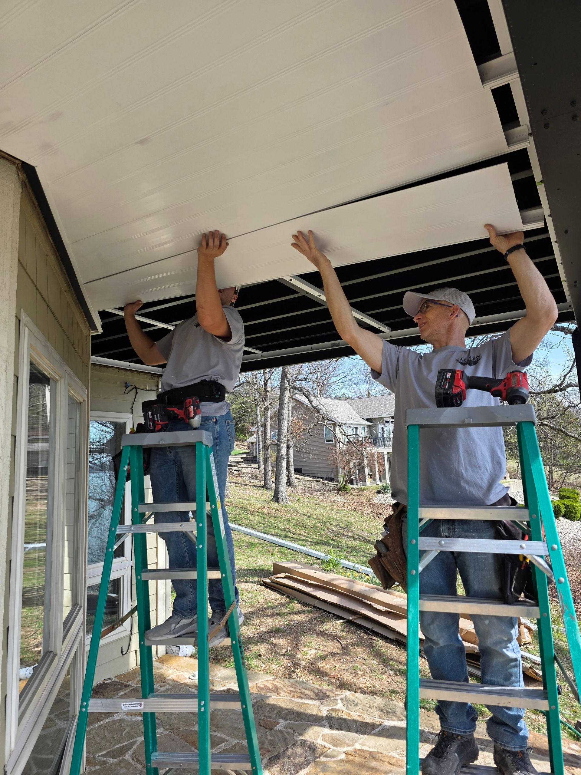 Two workers on ladders installing a white ceiling panel under a porch.