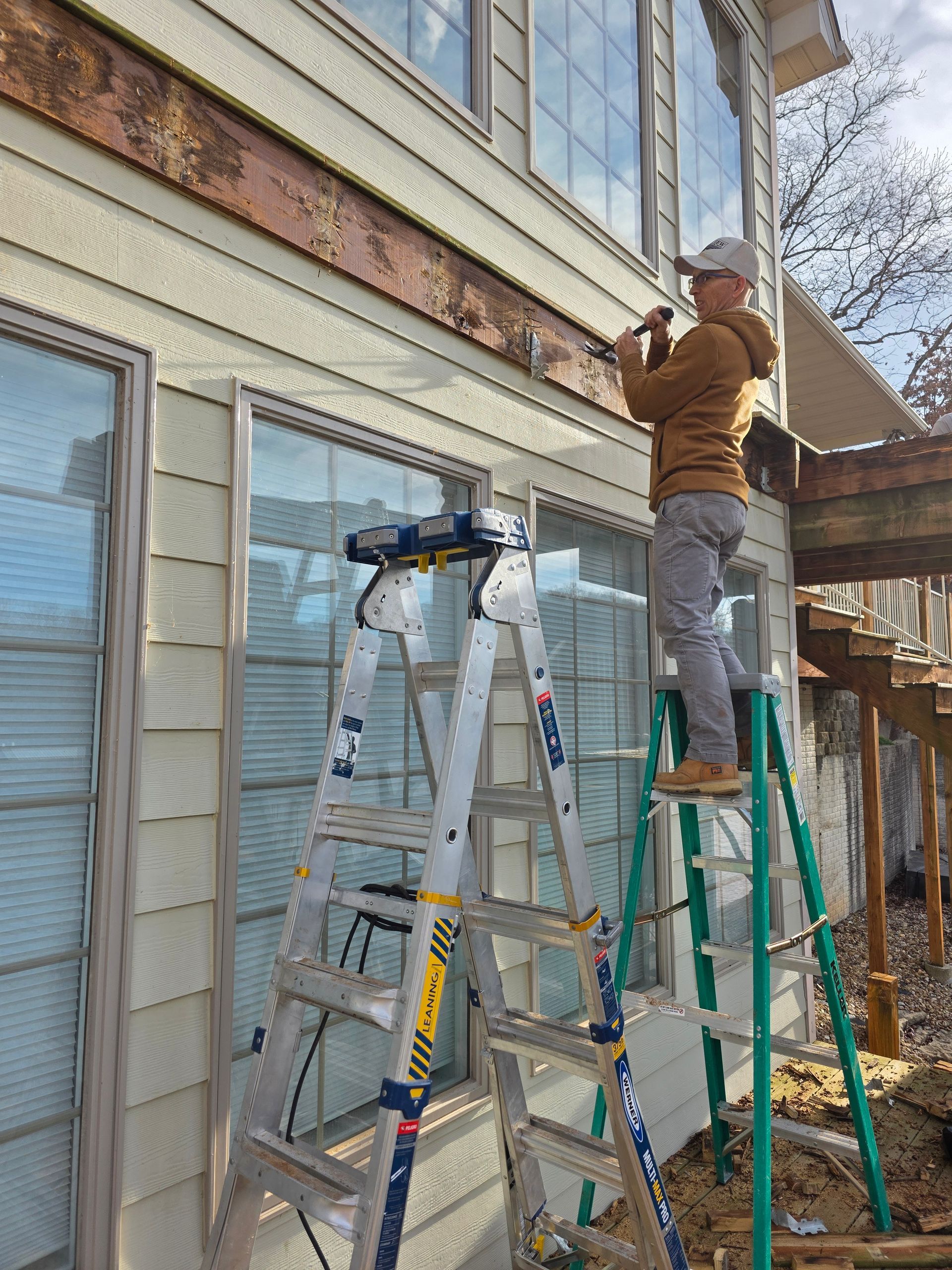 Worker on a ladder repairing exterior trim beside a house, with another ladder nearby.