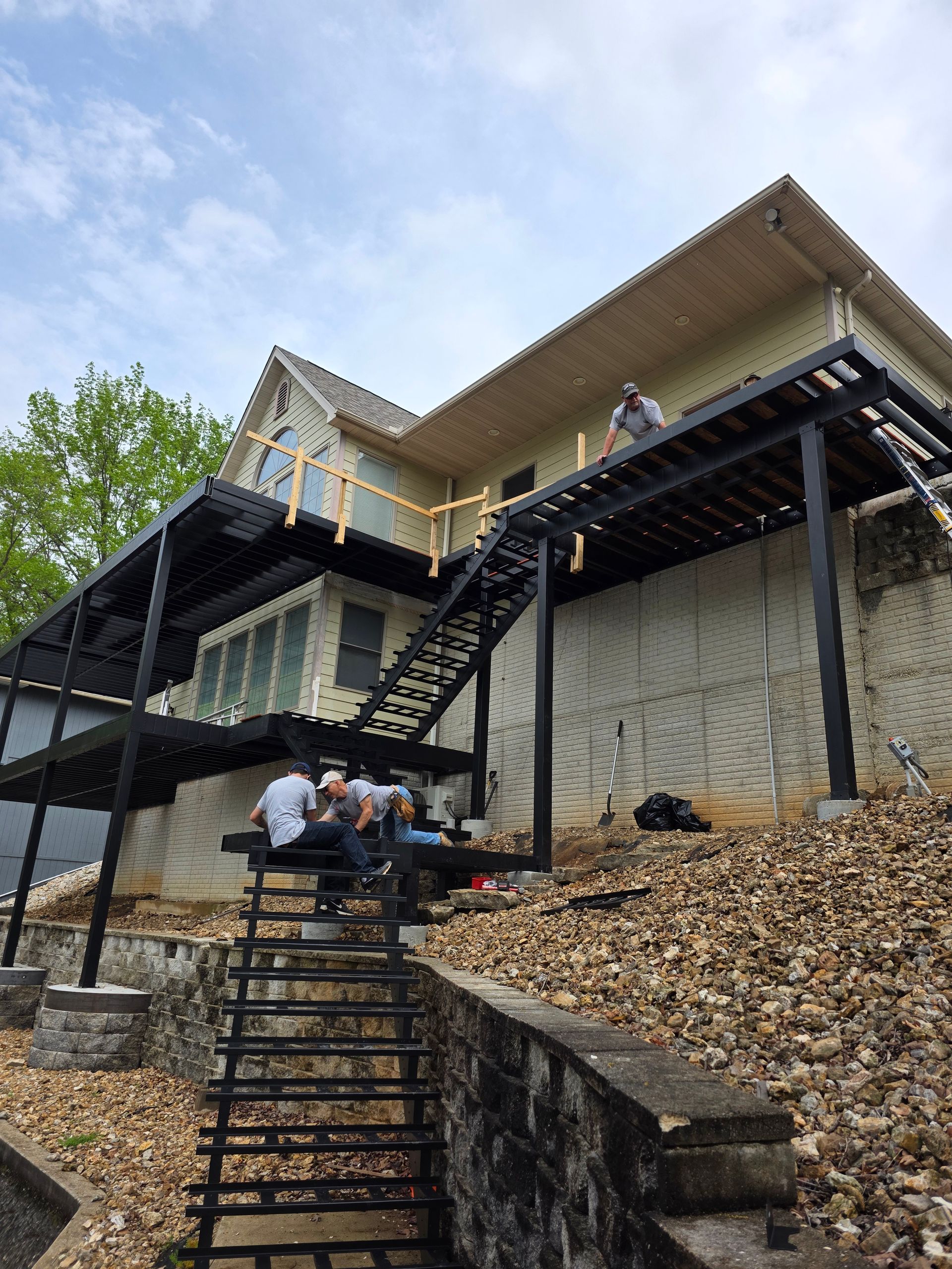 Two people sitting on steps beneath a modern house on a steep hillside with black railings and stone walls