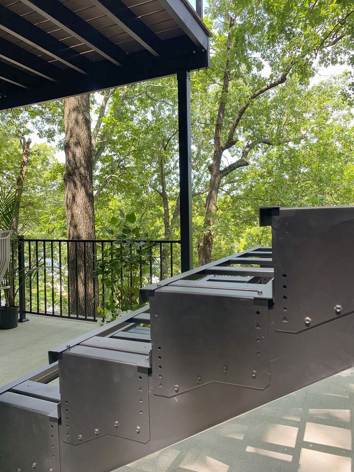 Outdoor stairs with metal frame, leading up to a deck overlooking trees. Gray and black.