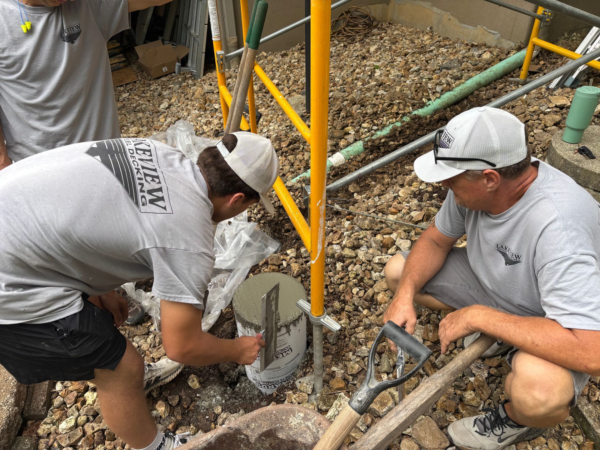 Two workers in gray shirts crouch beside a yellow scaffold, working in gravel with hand tools.