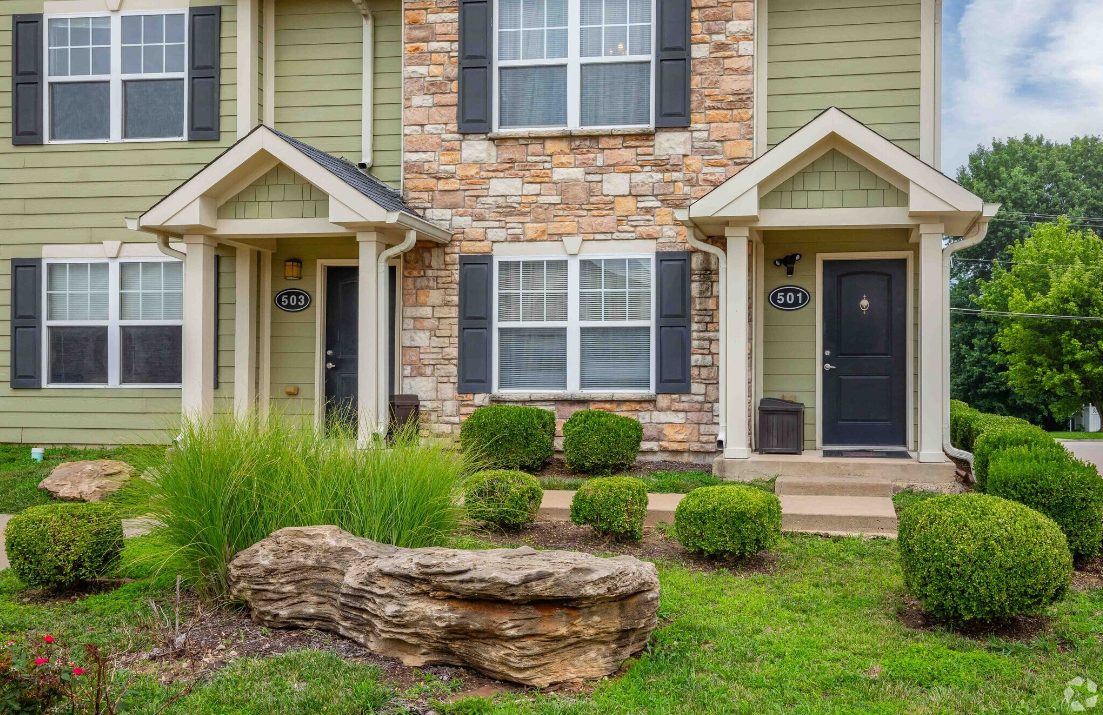 The front of a green and stone apartment building with a large rock in front of it.