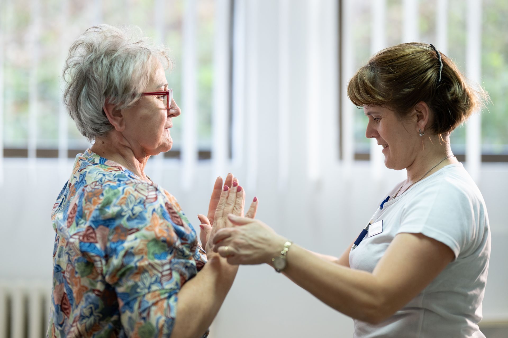 Older woman getting physical therapy for her hands from a younger therapist; indoor setting, light background.