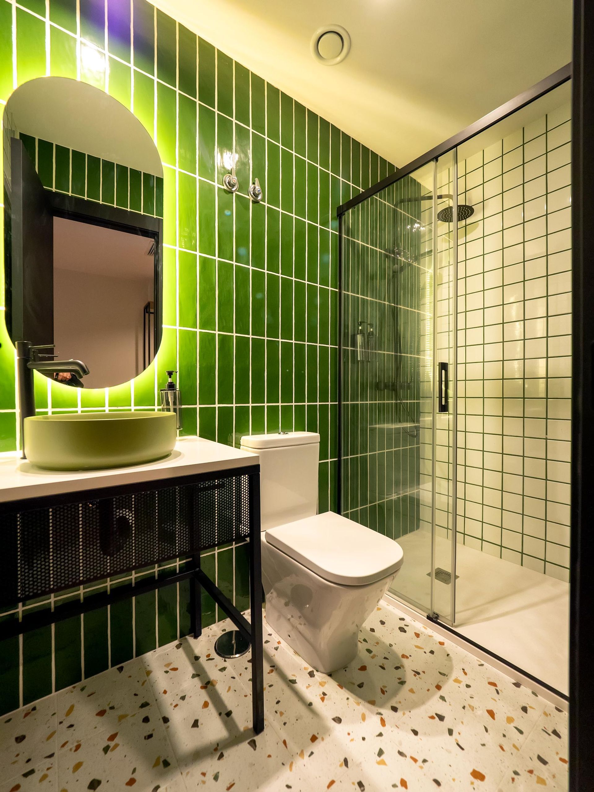 A bathroom with green tiles , a toilet , sink and shower.