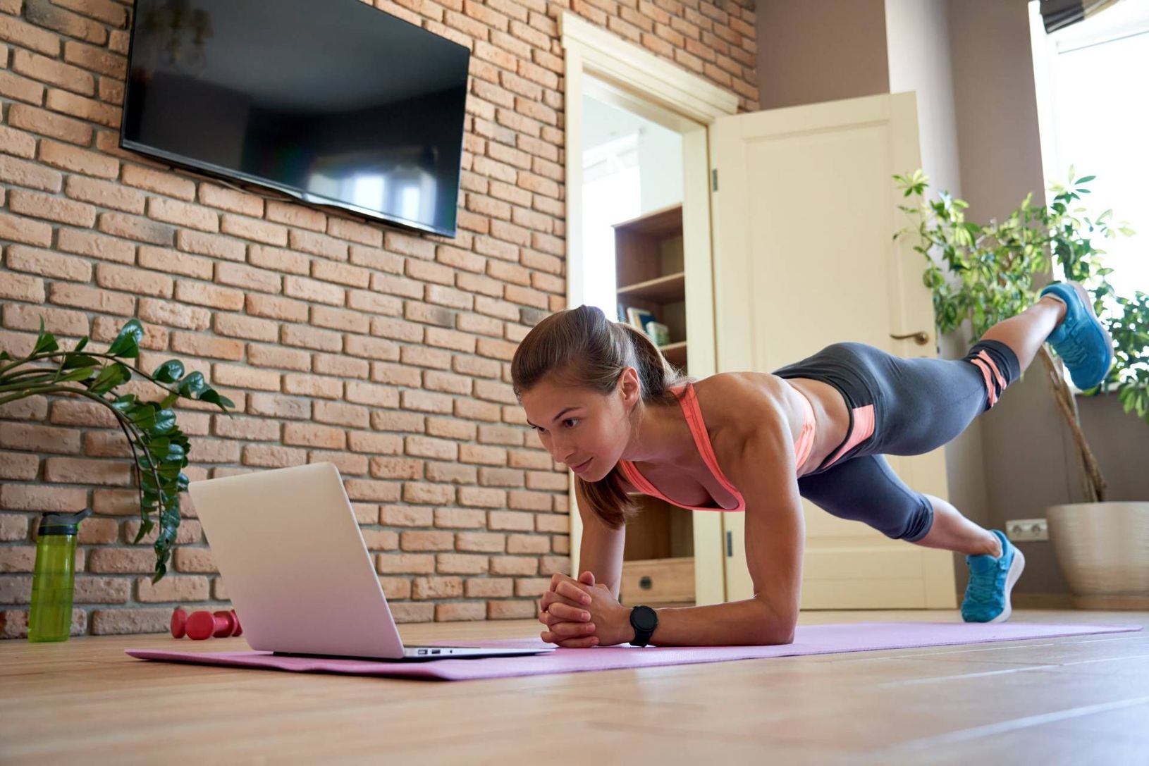 Woman doing a plank workout on a laptop in a living room with a brick wall.