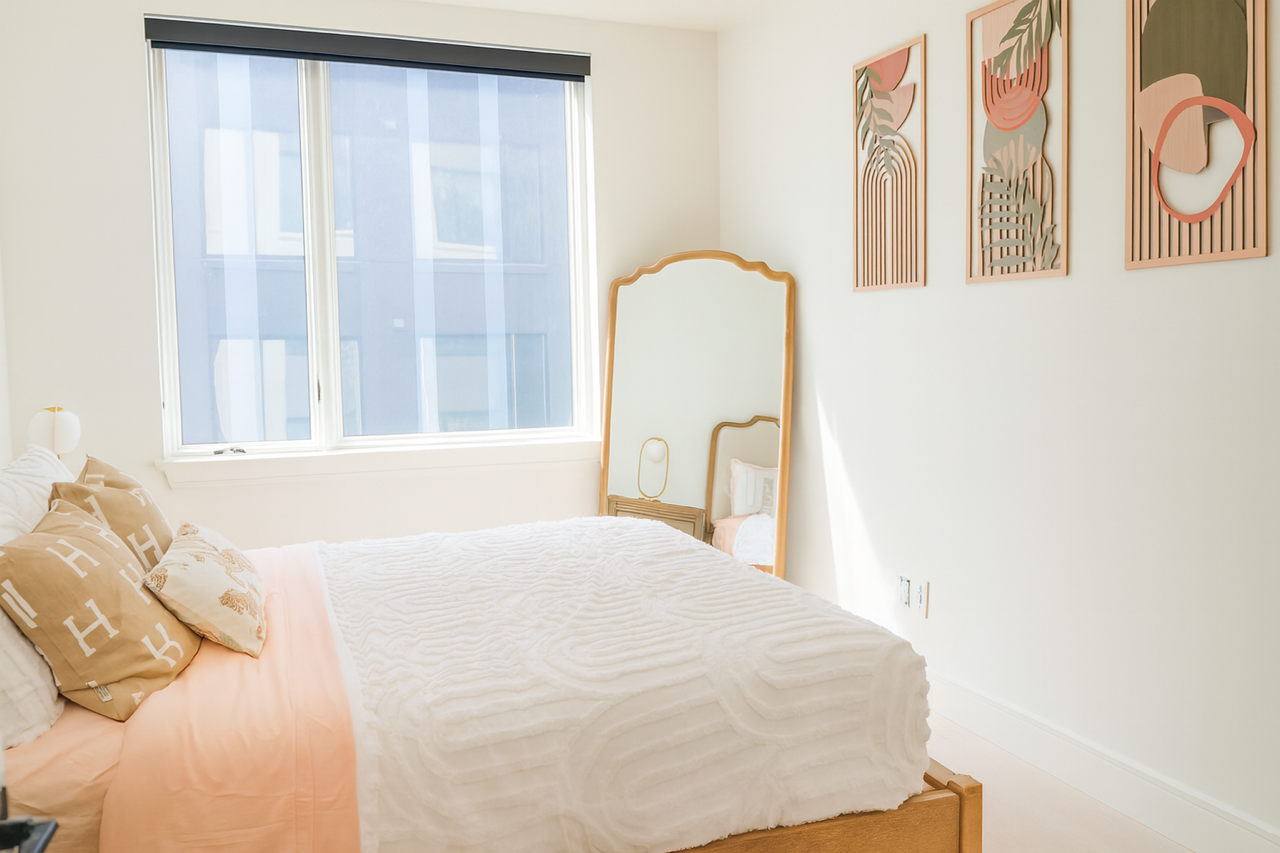 A bright bedroom with a bed featuring peach bedding, a white textured quilt, and a tall floor mirror near a large window.