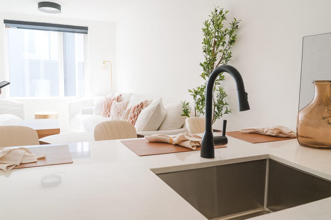 A bright kitchen island with a black faucet, sink, and place settings, with a living room visible in the background.