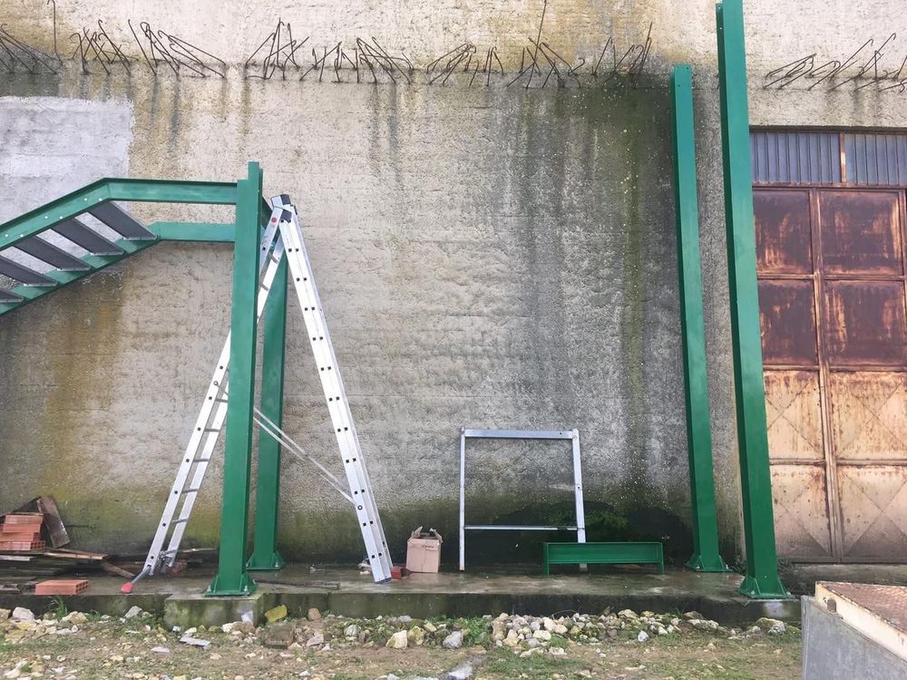 Struttura metallica verde in costruzione contro un muro di cemento strutturato, con una scala e degli attrezzi.