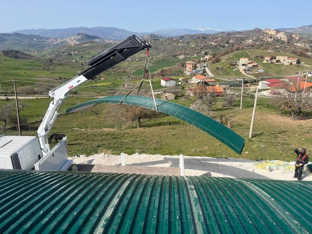 Gru che solleva un pannello di metallo curvo verde sopra un tetto simile in un ambiente rurale con montagne.