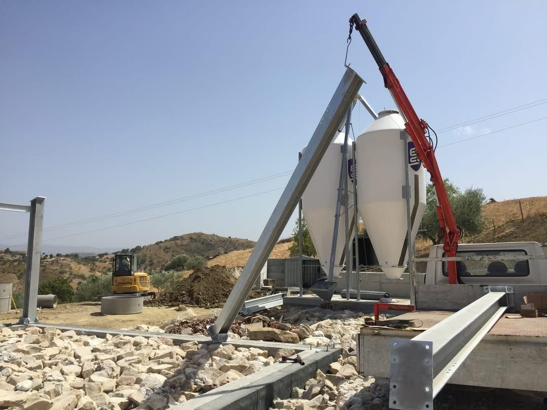Una gru solleva due silos bianchi a forma di cono durante la costruzione in un ambiente esterno.