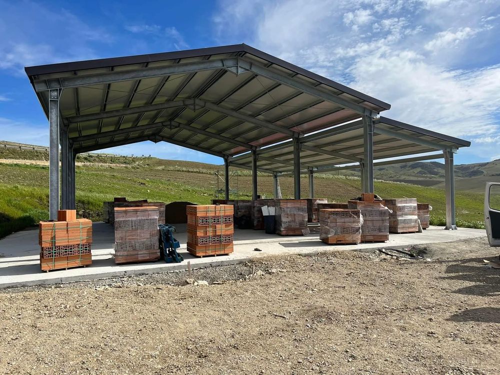 Un grande capannone aperto protegge pile di mattoni su una piattaforma di cemento. In un campo con verdi colline.
