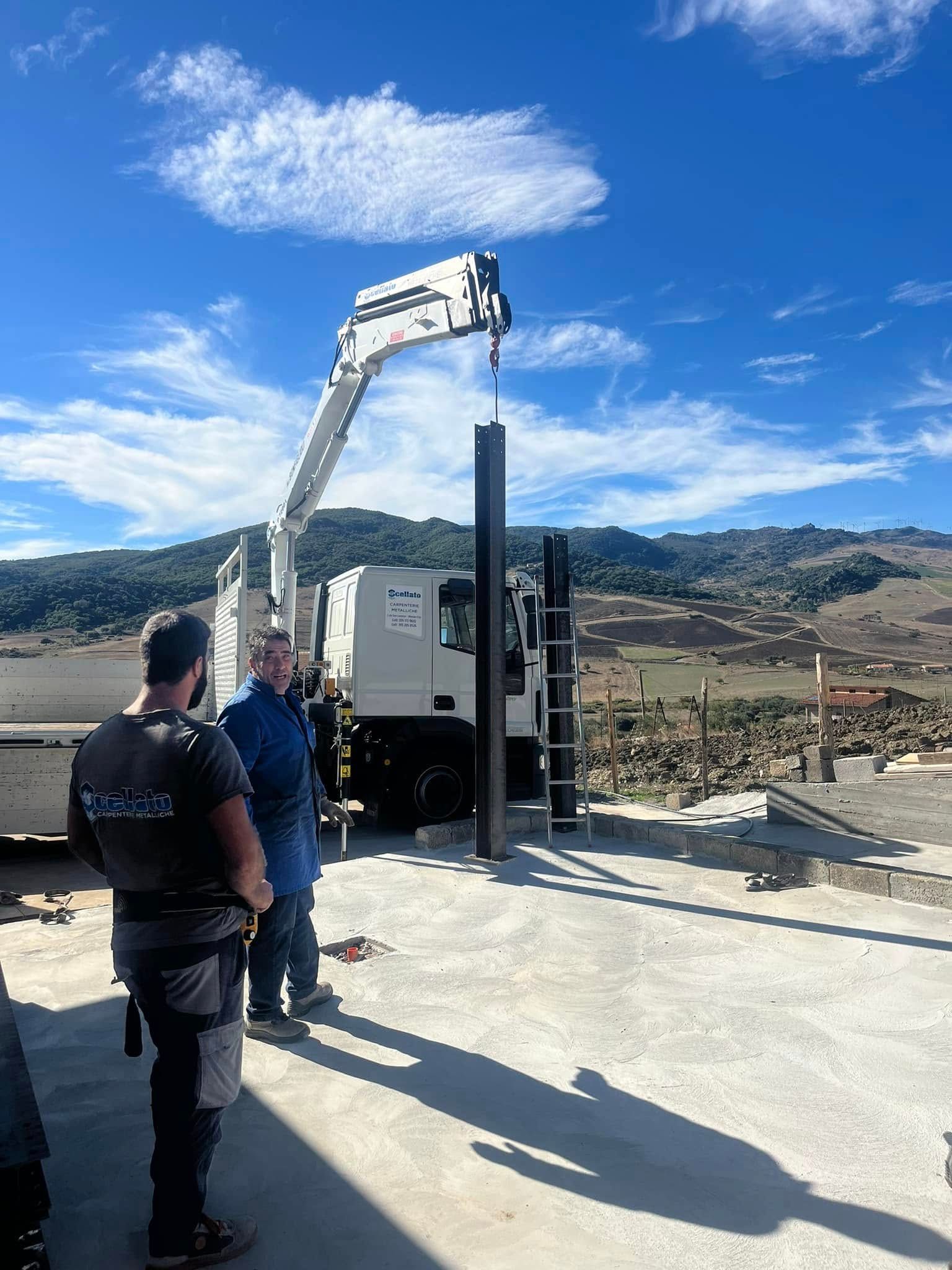 Operai edili accanto a una gru che solleva una trave metallica su un tetto con vista sulle montagne.