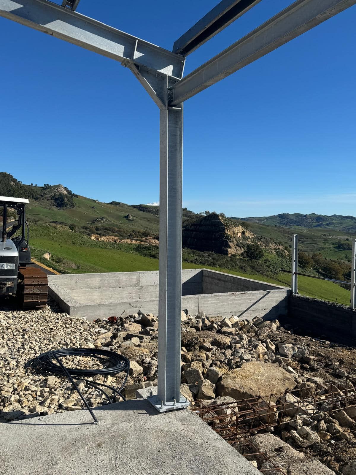 Cantiere con struttura in acciaio sullo sfondo di verdi colline e cielo azzurro.