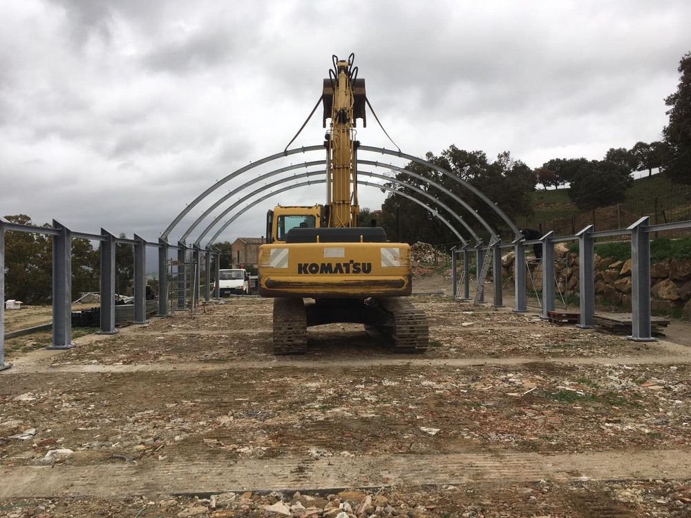 Escavatore che solleva una struttura metallica ad arco e la posiziona in un cantiere. Cielo nuvoloso.