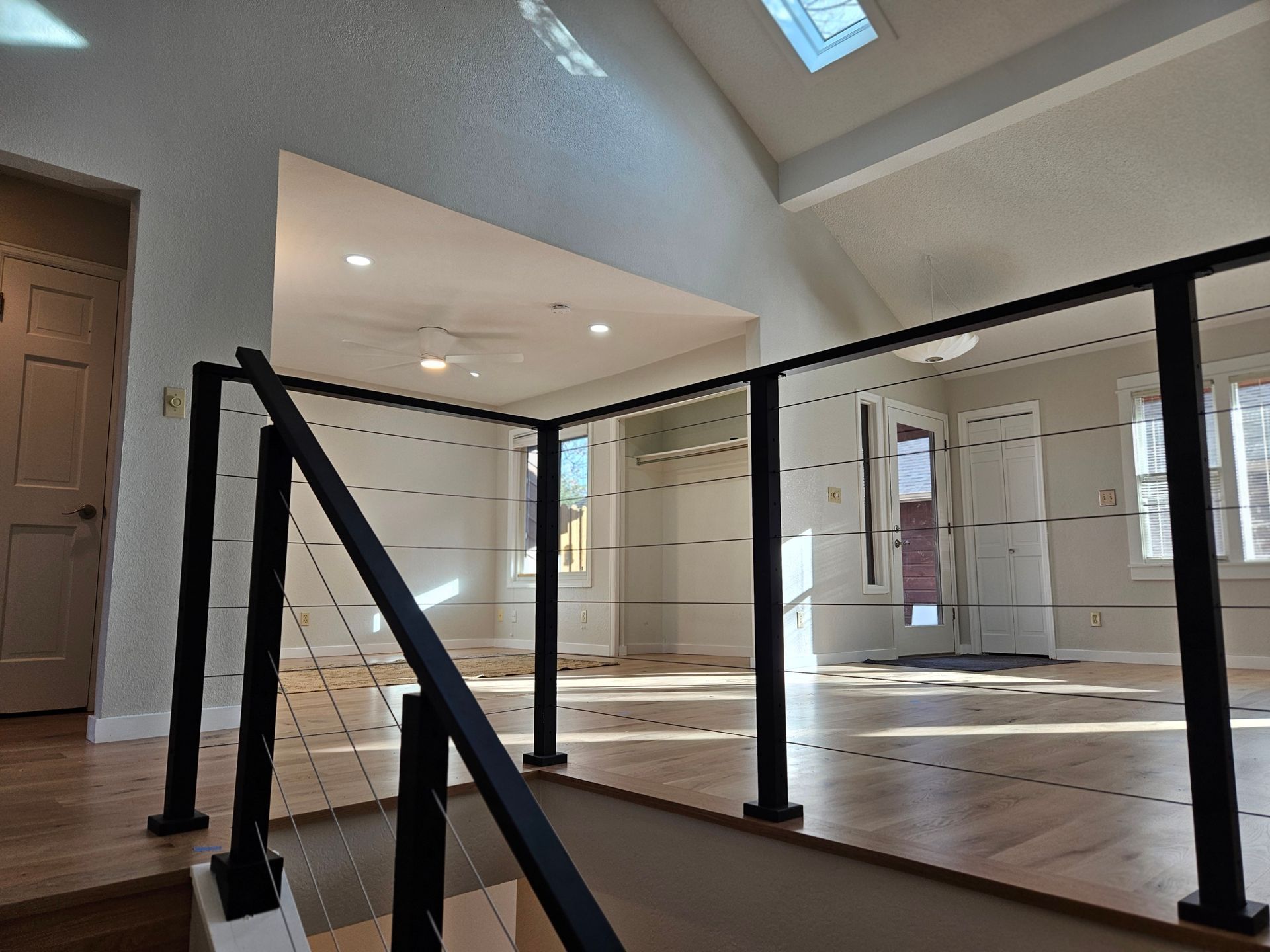 Interior view: A modern stair railing with black posts and clear panels overlooks a spacious room with wood floors and a high ceiling.