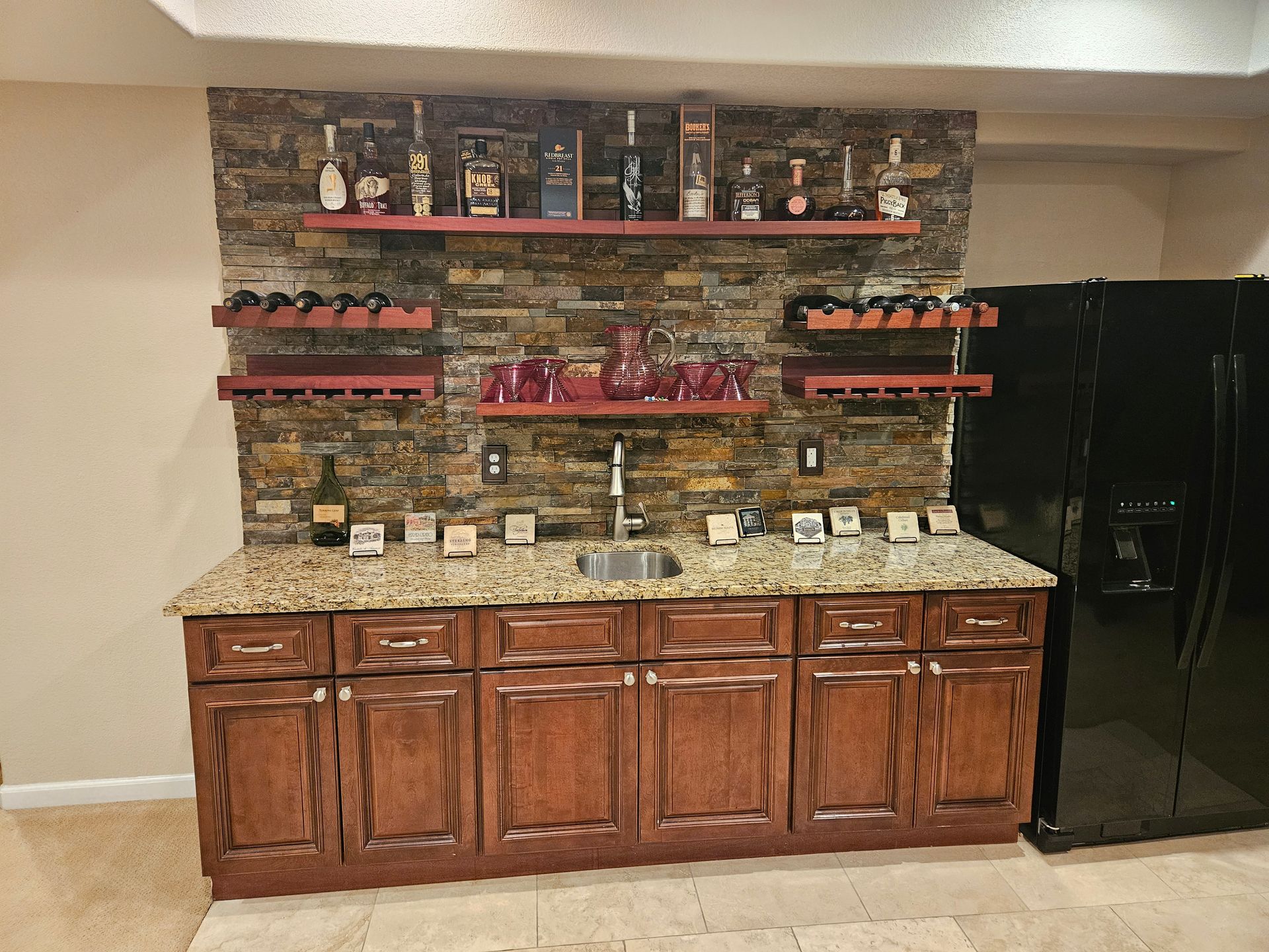 Home bar with brown cabinets, stone backsplash, shelves with bottles, and a sink.