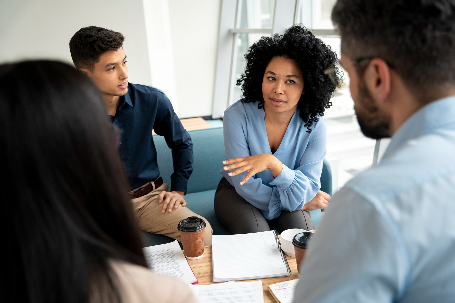 Four people in a meeting, one woman gesturing, papers and coffee on the table.