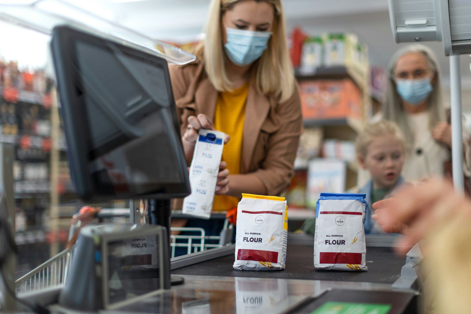 A woman wearing a mask is standing at a cash register in a grocery store.