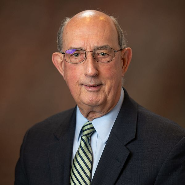 Man in a suit and tie, wearing glasses, looking at the camera, with a neutral expression, against a brown backdrop.