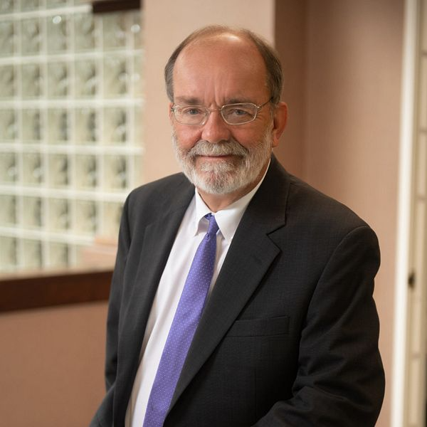 Man in suit, glasses, and tie smiles in an office setting, next to a block-glass wall.