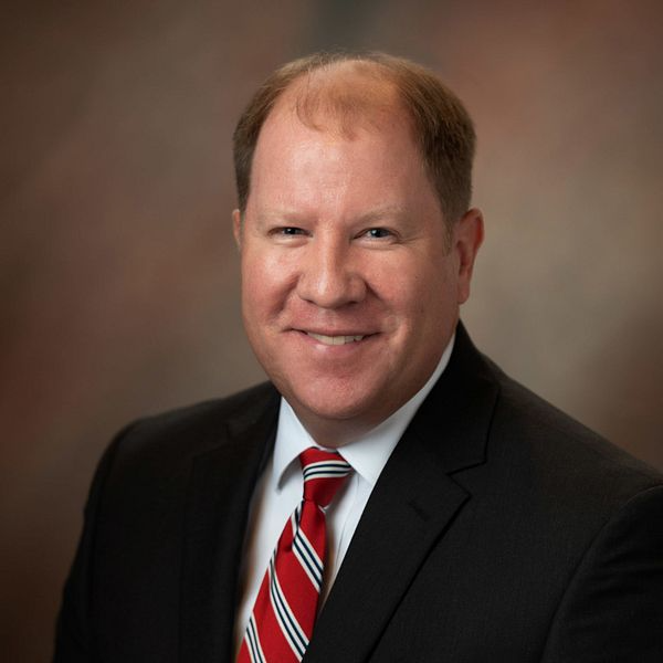 Man in suit and tie smiles at the camera, against a brown background.