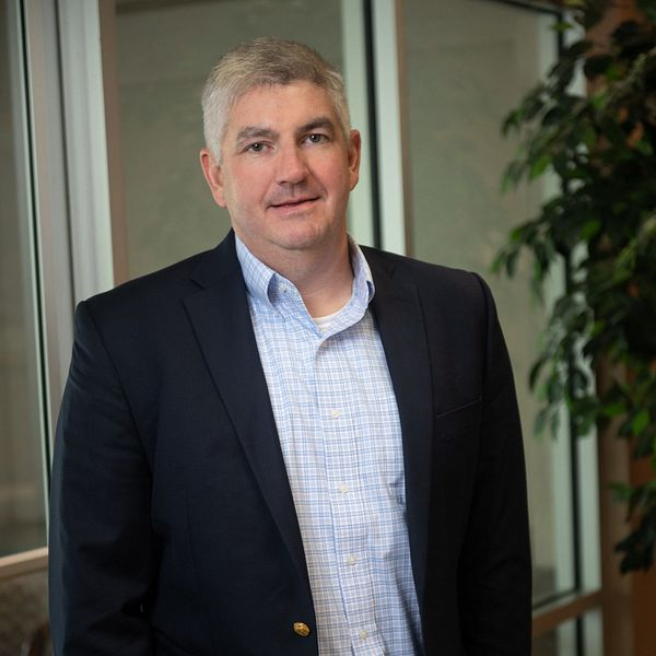 Man in navy blazer and patterned shirt standing by a window, smiling slightly.