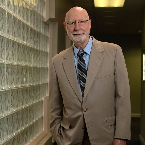 Man in beige suit and tie, leaning against a glass block wall in a hallway, smiling.