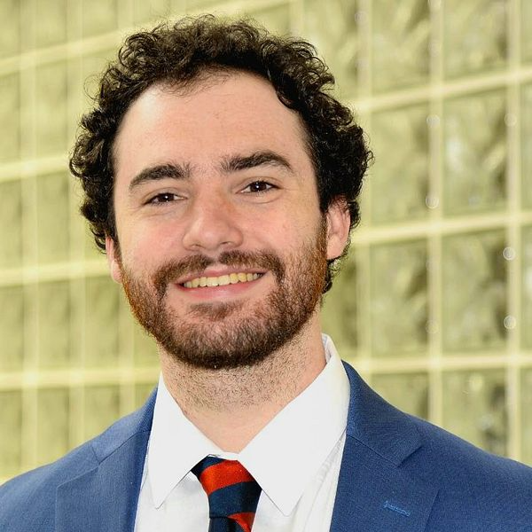 Man in blue suit, smiling, red and blue striped tie, brown curly hair, and beard.