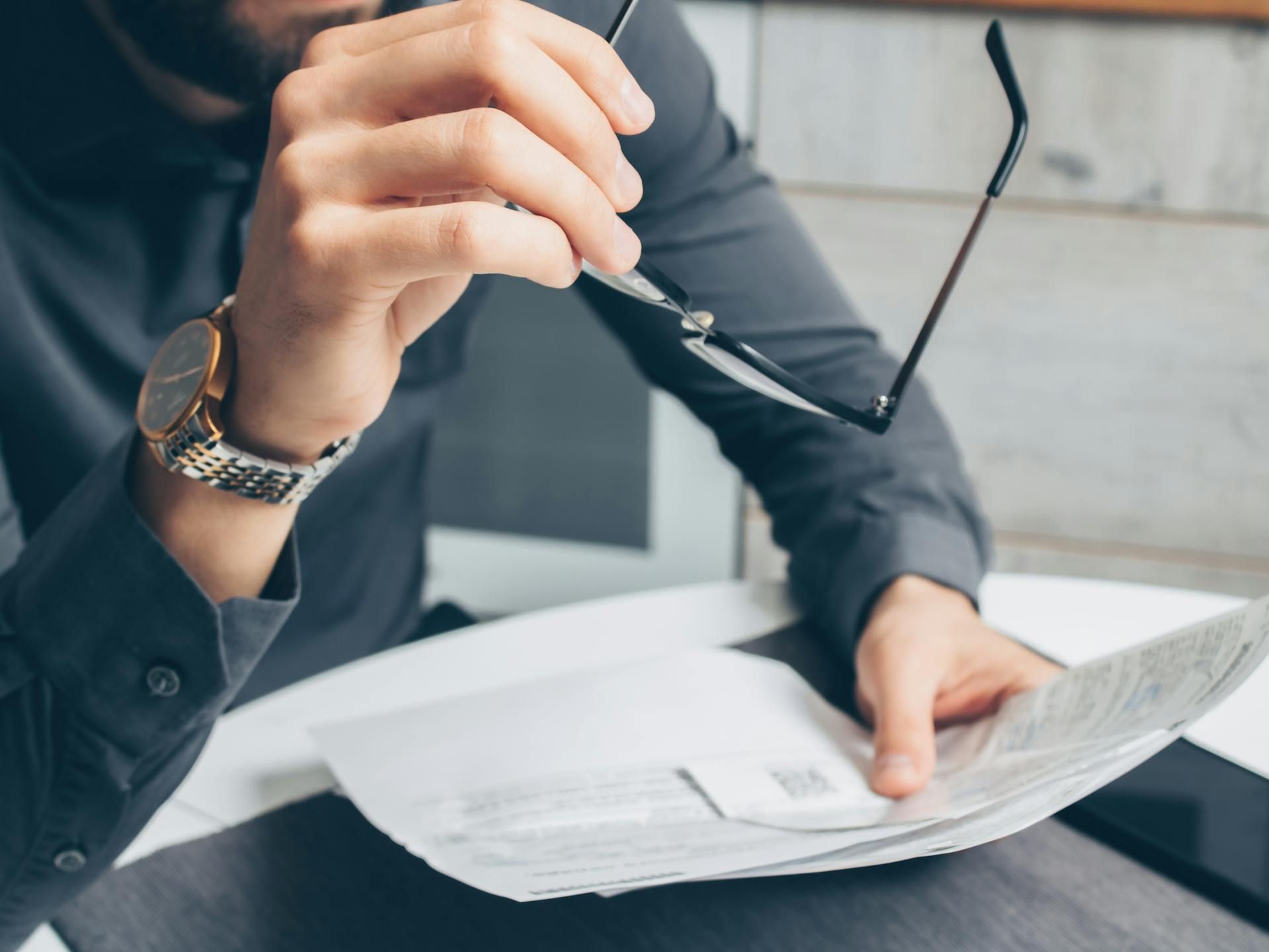 Man holding glasses, reviewing documents; wearing a watch and dark button-down shirt.