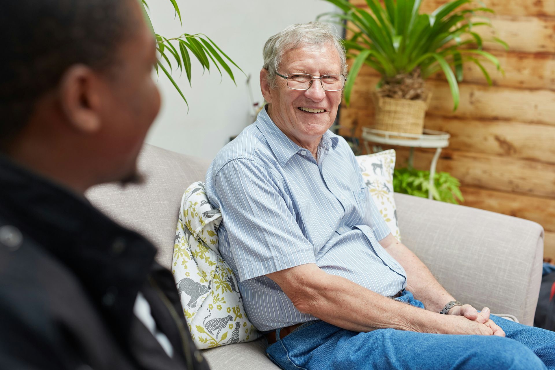 Man smiles at another person while seated on a sofa with plants in the background.